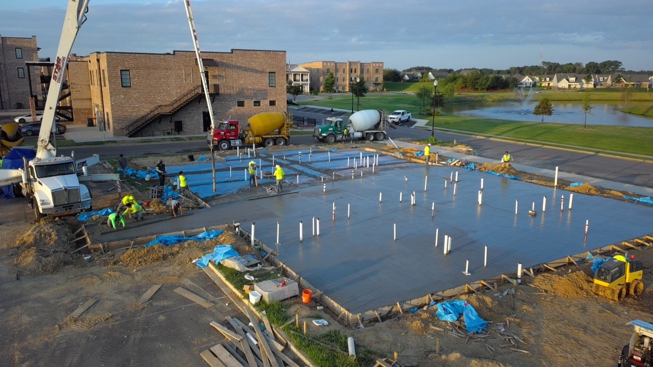 Aerial view of an active concrete slab pour at a multi-family development, with a pump truck and mixer truck on site and crew members in yellow safety vests finishing the fresh slab